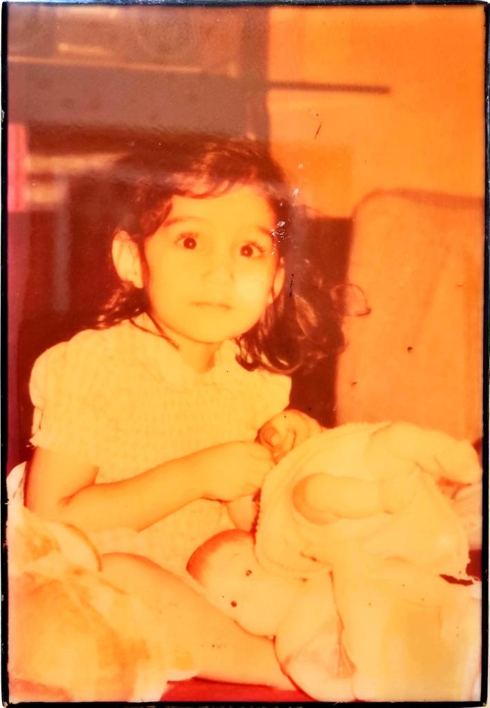 Sepia-toned image of toddler Mariyam sitting and smiling at the camera. She wears a frilly dress and her curly hair is in a little ponytail.