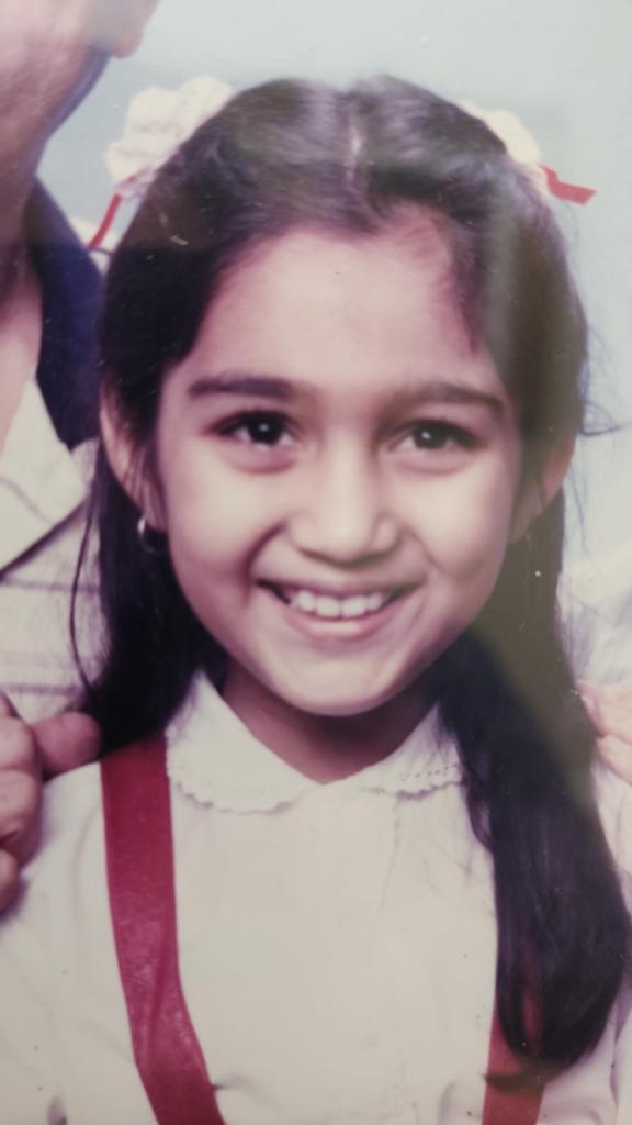 Seven year old Mariyam smiles in her first family portrait with her parents (cropped out for their privacy). She wears a white blouse and red pinafore dress. 